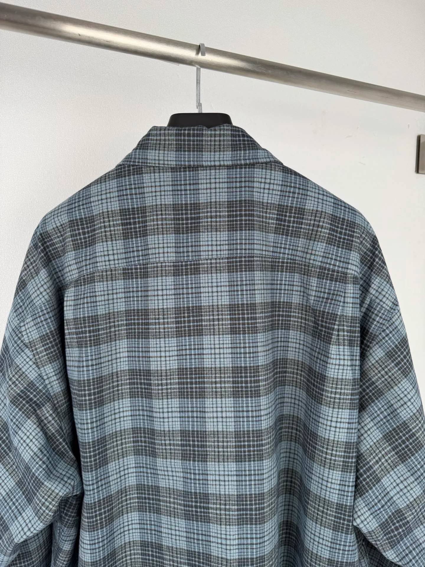 BALENCIAGA/balenciaga blue and gray plaid zipper pocket logo quilted jacket combines a three-layer structure of plaid woven fabric, filling cotton, and lining, and integrates plaid alignment technology and quilted garment production standards. There are multiple process difficulties in the whole process. The surface layer of the body is made of blue-gray Retro plaid fabric, and the cutting machine production strictly follows the global grid standards. The arrangement direction of the cutting pieces is fixed and cannot be changed at will. The horizontal and vertical lines of shoulder seams, side seams, and zipper placket joints must be consistent and coherent. The left chest patch pocket is cut independently, and the bag check pattern must be aligned with the front reference pattern. Slight deviations will destroy the visual integrity and directly increase fabric loss and cutting time. The most difficult part of the style is the three-layer composite sewing. The outer plaid fabric, filling cotton, and lining fabric have different physical properties, and the preshrinking processing parameters need to be adjusted separately. Pressure and temperature control are crucial during lamination operations. Once the parameters are out of balance, it can easily cause fabric stretch and distortion, plaid dislocation, and wrinkles and bulges in garments. At the same time, it is necessary to reasonably plan the quilting line to avoid the visual beauty of the grid pattern from being cut by the lines on the basis of firmly fixing the cotton layer and preventing cotton from running away. The placket adopts a centered zipper structure. It is difficult to sew the zipper tape and the quilted panels. It is required that the width of the left and right plackets be equal, the zipper routing is straight and smooth, and there is no lag in opening and closing. When the shirt lapel is filled with cotton, the thickness of the cotton needs to be controlled to prevent the collar from becoming bloated and unnaturally folded. The metal nameplate of the chest pocket is sewn at fixed points to avoid pulling the fabric during the operation and ensure that the plaid pattern is smooth and not deformed. The quilted material increases the overall thickness of the fabric. When sewing, you need to continuously adjust the presser foot pressure and stitch length to adapt to changes in sandwich thickness. The low-temperature and gentle ironing process is used for post-finishing to prevent the plaid from fading and the cotton layer from clumping due to high temperatures. From the precise alignment of cutting pieces, three-layer fabric pretreatment, composite quilting to garment finalization inspection, the processes are interlocking and the error tolerance rate is extremely low, testing the accuracy of plate making, equipment control and turning technology. Size:XS S M L